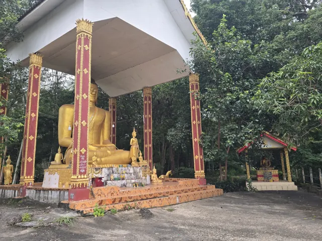 ພຣະພຸດທະຮູບສີທອງພາຍໃນສາລາ ລ້ອມຮອບດ້ວຍຮົ່ມໄມ້.
